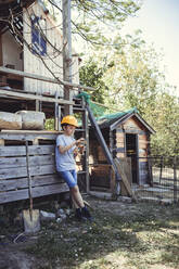 Boy in yellow hardhat calculating on toy while leaning on rabbit hutch in back yard - HMEF01278