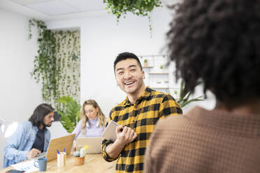 Smiling young businessman with digital tablet looking at female coworker while discussing in office - JCCMF02693