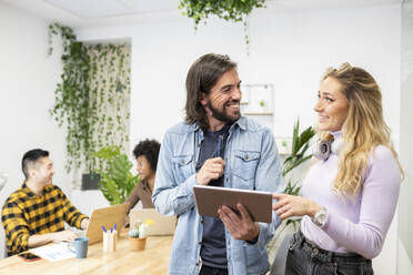 Cheerful male and female colleague looking at each other while discussing over digital tablet in office - JCCMF02700