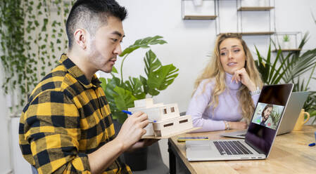 Male architect explaining house model though video call in office - JCCMF02706