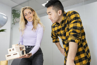 Young businessman discussing with female colleague holding model house in office - JCCMF02718