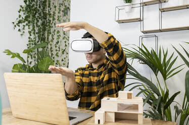 Young male professional wearing virtual reality simulator gesturing while sitting at desk in office - JCCMF02725