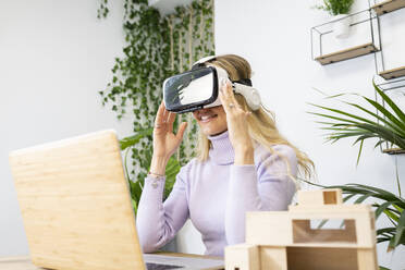 Lächelnde blonde Geschäftsfrau, die einen Virtual-Reality-Simulator trägt und am Schreibtisch im Büro sitzt - JCCMF02727