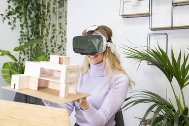 Lächelnde weibliche Fachkraft, die einen Virtual-Reality-Simulator trägt und ein Hausmodell im Büro hält - JCCMF02729