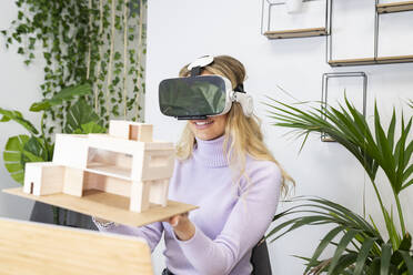 Smiling female professional wearing virtual reality simulator holding house model in office - JCCMF02729