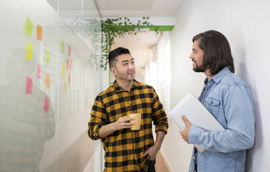 Male professional looking at colleague standing with laptop discussing while standing in office corridor - JCCMF02747