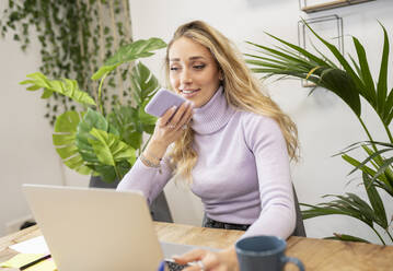 Smiling blond businesswoman talking on mobile phone though speaker in office - JCCMF02770