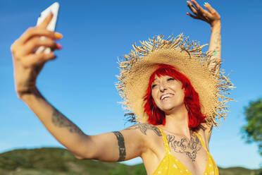 Cheerful tattooed woman in sun hat taking selfie on sunny day - MRRF01220