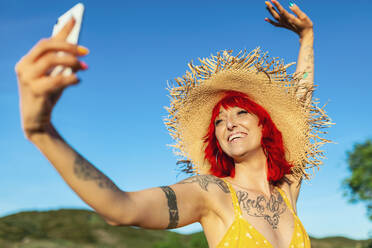 Fröhliche tätowierte Frau mit Sonnenhut, die an einem sonnigen Tag ein Selfie macht - MRRF01220