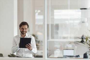 Lächelnder männlicher Unternehmer, der ein digitales Tablet hält, während er im Büro steht - GUSF05950