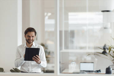 Smiling male entrepreneur holding digital tablet while standing in office - GUSF05950