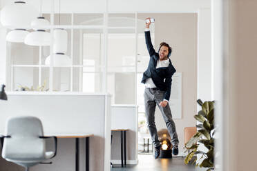 Geschäftsmann mit Mobiltelefon, der beim Springen im Büro jubelt - GUSF05978