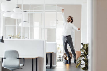 Excited businessman jumping with hand raised in office - GUSF05979