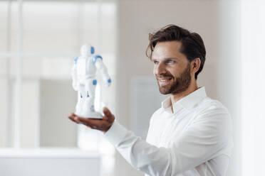 Männlicher Unternehmer, der einen Roboter hält, während er im Büro steht - GUSF05981