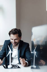 Lächelnder männlicher Unternehmer, der ein Windradmodell im Büro betrachtet - GUSF05995