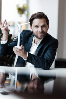 Smiling businessman gesturing while holding wind turbine model in office - GUSF05998