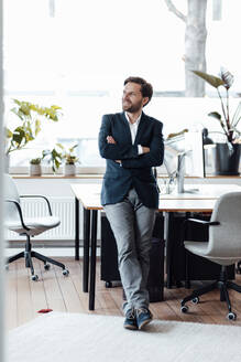 Businessman with arms crossed leaning on desk in office - GUSF06002