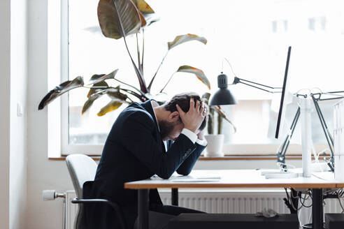 Male entrepreneur with head in hands sitting by desk at office - GUSF06003