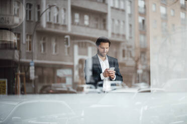 Männlicher Unternehmer, der ein Mobiltelefon benutzt, während er am Fenster steht - GUSF06016