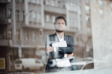 Nachdenklicher männlicher Unternehmer, der ein digitales Tablet hält und durch ein Bürofenster schaut - GUSF06019