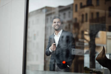 Männlicher Unternehmer, der ein digitales Tablet hält, während er am Bürofenster steht - GUSF06021