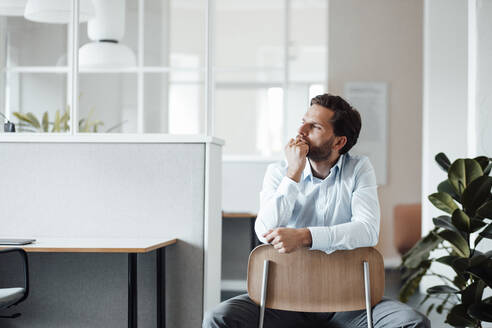 Nachdenklicher männlicher Unternehmer, der wegschaut, während er auf einem Stuhl im Büro sitzt - GUSF06034