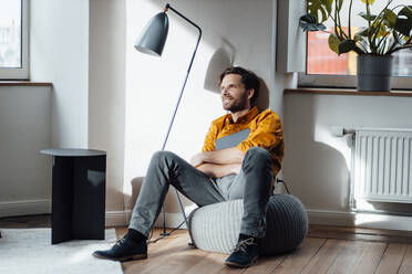 Nachdenklicher männlicher Unternehmer, der ein digitales Tablet hält und auf einem Hocker sitzt - GUSF06048