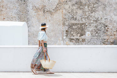 Frau mit Hut geht an einem sonnigen Tag auf einem Fußweg an einer Stützmauer spazieren - JCMF01974