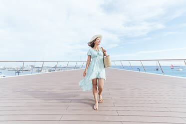 Lächelnde Frau mit Hut, die wegschaut, während sie auf dem Pier spazieren geht - JCMF01981