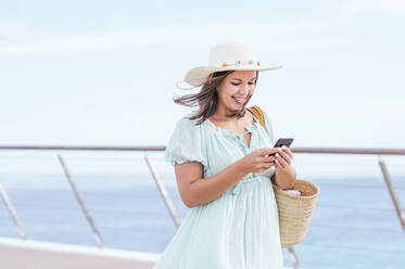 Lächelnde reife Frau mit Hut und Smartphone am Geländer - JCMF01983