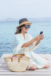 Smiling mature woman wearing hat taking selfie through smart phone while sitting on jetty - JCMF01986