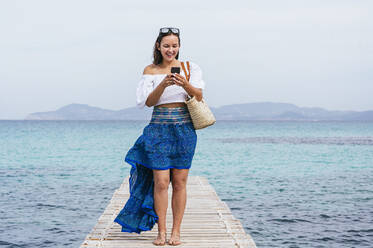 Smiling woman using smart phone while standing on jetty - JCMF01990