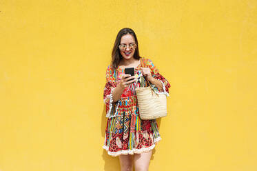 Lächelnde Frau mit Smartphone vor einer gelben Wand an einem sonnigen Tag - JCMF01994