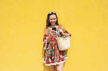 Lächelnde reife Frau, die eine Textnachricht über ihr Mobiltelefon sendet, während sie vor einer gelben Wand an einem sonnigen Tag steht - JCMF01995