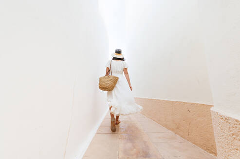 Mature woman wearing hat walking on footpath amidst walls - JCMF01998