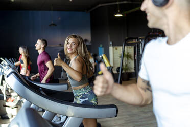 Smiling male and female athletes gesturing thumbs up to each other while exercising on treadmill at gym - MPPF01755