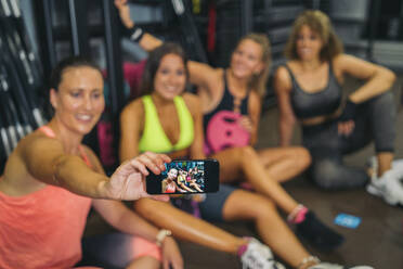 Lächelnde Frau nimmt Selfie mit weiblichen Freunden durch Smartphone im Fitnessstudio - MPPF01759
