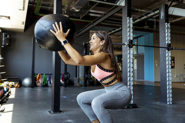 Smiling female athlete crouching while exercising with fitness ball at gym - MPPF01762