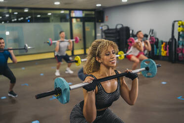 Curly haired female athlete lifting barbell while exercising with friends in gym - MPPF01804