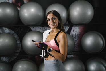 Lächelnde Sportlerin, die ihr Mobiltelefon an Fitnessbällen im Fitnessstudio hält - MPPF01815
