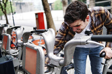 Smiling young man unlocking bike at station - ASGF00427