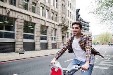 Lächelnder Mann mit kariertem Hemd, der auf einer Straße in der Stadt Rad fährt - ASGF00430