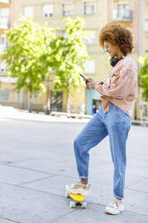 Junge Frau benutzt ihr Smartphone, während sie auf einem Skateboard auf dem Gehweg steht - IFRF00751