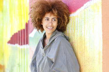 Smiling young woman looking away while leaning on colorful wall - IFRF00766