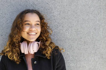 Smiling young woman with headphones contemplating in front of wall - IFRF00780