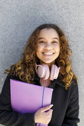 Smiling young woman with headphones holding file against wall - IFRF00781