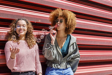 Young female friends playing with bubbles in front of corrugated wall on sunny day - IFRF00795