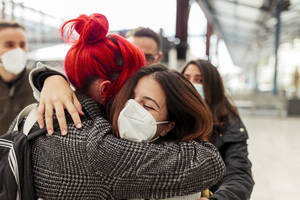 Female friends in face masks embracing at railroad station during COVID-19 - MRRF01231