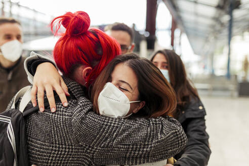 Female friends in face masks embracing at railroad station during COVID-19 - MRRF01231