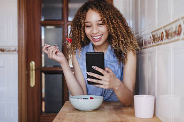 Lächelnde junge Frau benutzt ihr Smartphone beim Frühstück in der Küche - EBBF03826